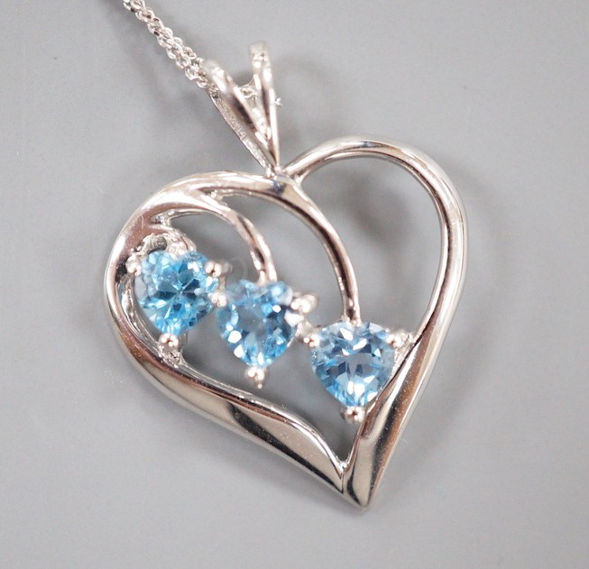 A modern 9ct white gold and three stone blue topaz set stylised heart shaped, pendant, 25mm, on a 375 fine link chain, 40cm, gross weight 2.7 grams.